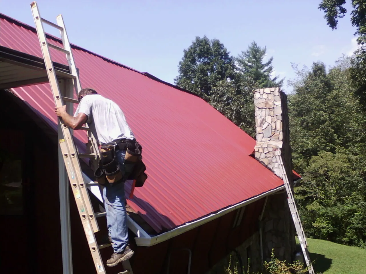 Roofer on a ladder beside a red metal roof and stone chimney — Soffit & Fascia Repair by Roofing Brothers in Grove City
